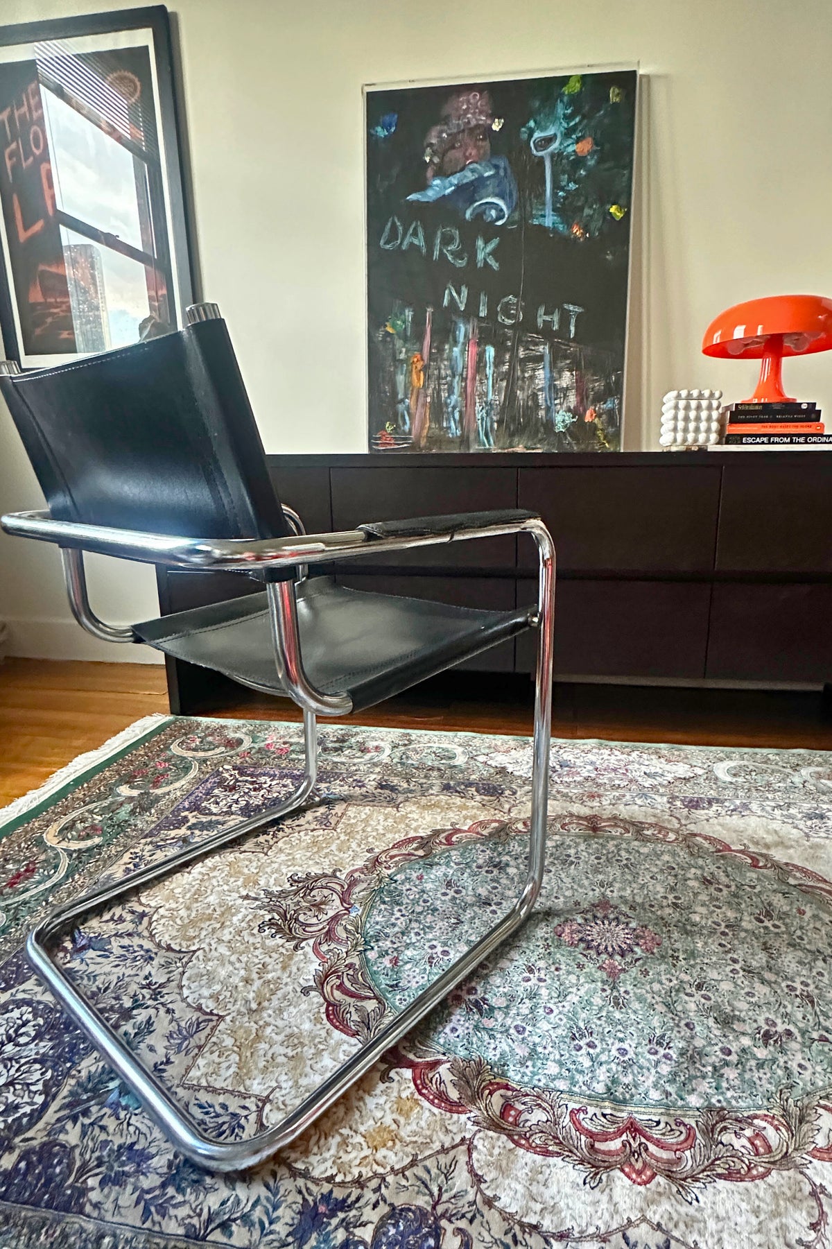 Modern chair on a patterned rug with a colorful painting and orange lamp in the background.