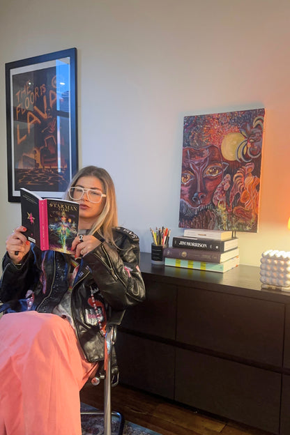 Person reading a book in a room with a painting and books on a shelf.